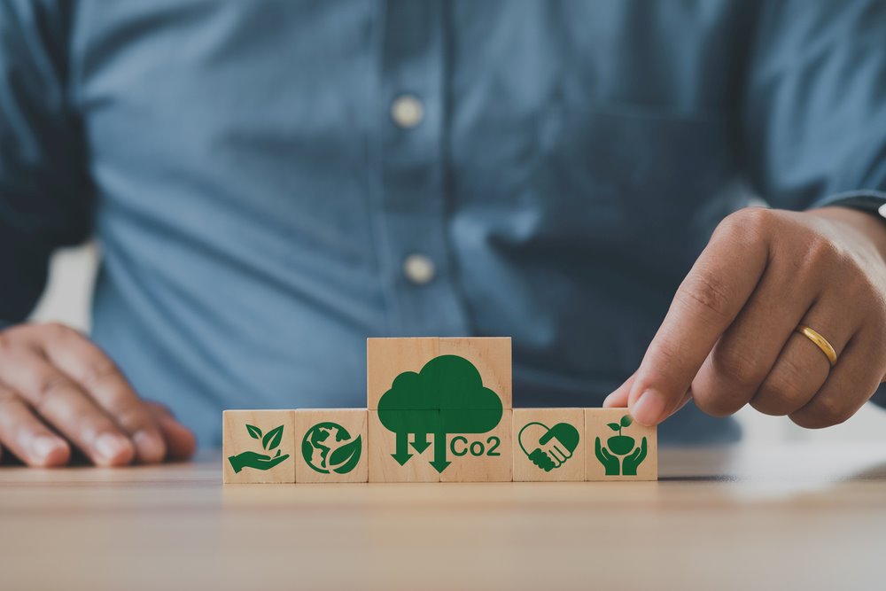 Image of a man's hand placing wooden cubes with a CO2 emission reduction icon, featuring 411 Business Law and highlighting 'Assessing Your Regulatory Environment,' ensuring it aligns with the page's context.